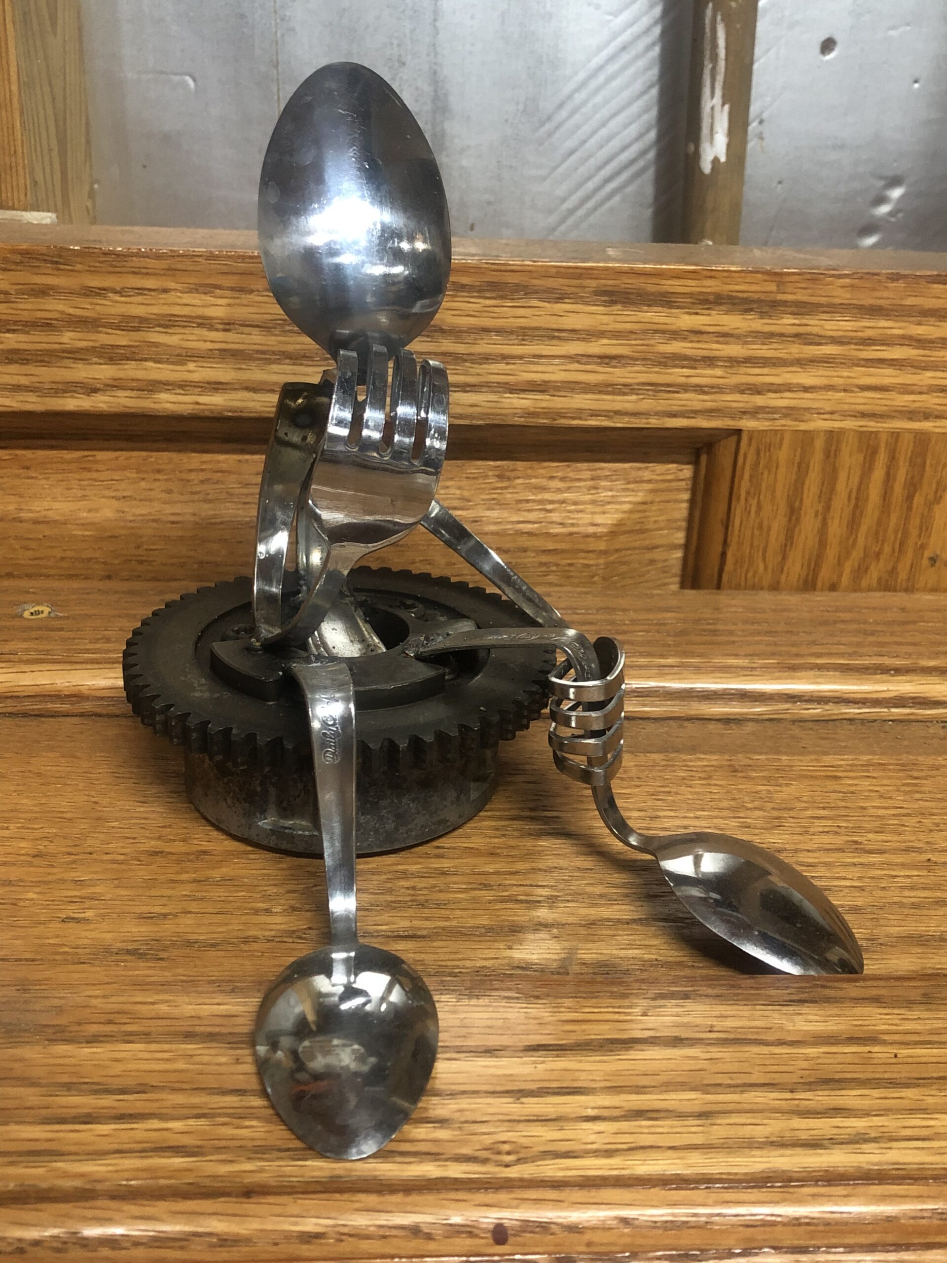 Spoons, forks, and gears assembled to make a "person" sitting in a "chair". Handmade by "Off The Cuff Metal Art"