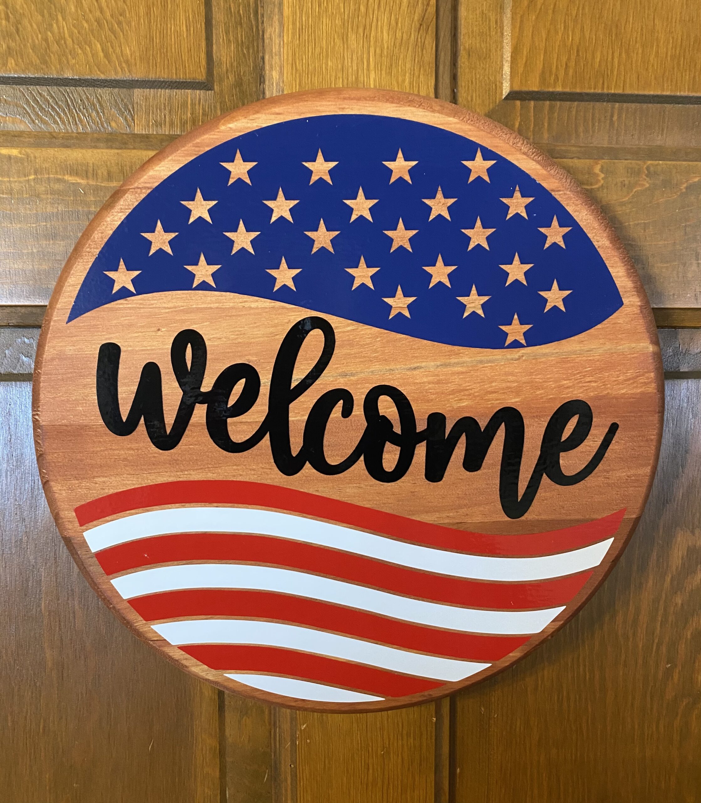 Wooden, round, sign that says "Welcome" and has a deconstructed American flag on it. 